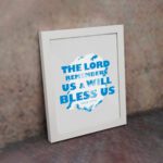 Wall art featuring the text The Lord Remembers Us And Will Bless Us in bold blue on a soft gray background. displayed on the wall in a white frame