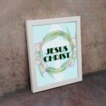 Infidu Jesus Christ Bible Wall Art with JESUS CHRIST text in a mint blue background surrounded by a floral wreath. displayed on the wall in a white frame