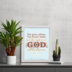 The Grass Withers Christian wall art with the verse Isaiah 40:8 in a modern font, featuring floral illustrations and a light blue background. displayed the table in a white frame