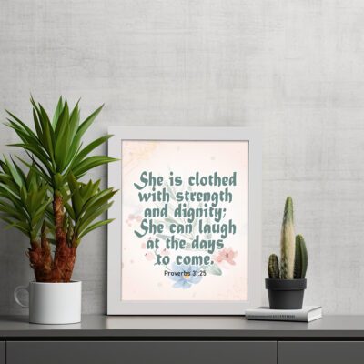 Wall art featuring Proverbs 31:25 with the quote in dark green text and floral decorations on a cream background. displayed on the table in a white frame