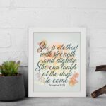 Christian wall art featuring the quote She is clothed with strength and dignity; she can laugh at the days to come from Proverbs 31:25, with an elegant design and pastel colors. displayed on the wall in a white frame