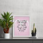 Proverbs 31:25 Christian wall art with She is clothed with strength and dignity on a soft pink background in an elegant script, framed. displayed on the table in a white frame