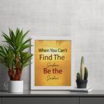 Infidu When You Can't Find The Sunshine Be The Sunshine wall art with a yellow background brown text and white frame on the table. perfect for inspiring positivity in any workspace.