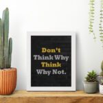 Infidu Don't Think Why, Think Why Not wall art with a black background yellow and white text, and a white frame on the table. Inspiring decor for home offices and workspaces.