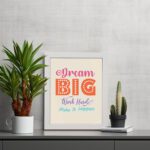 Infidu Dream Big Work Hard Make It Happen wall art with a light pink background and vibrant pink, orange, violet, and sky blue text and a white frame displayed on the table. Inspiring decor for home offices and workspaces.