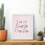 Infidu Take A Small Step Everyday wall art with a gray and light pink background black and red text and a white frame on the table. Inspiring decor for any workspace.