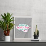 Infidu Take A Small Step Everyday wall art with a gray background and vibrant green, yellow, and pink text and white frame on the table. Inspiring decor for any workspace.