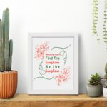 Infidu When You Can't Find The Sunshine Be The Sunshine wall art with a white background and red floral accents and white frame on the table. Inspiring decor for home offices.