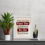 Infidu motivational wall art featuring the quote When You Can't Find the Sunshine Be the Sunshine on a white background with red text and a white frame on the table. perfect for office decor.