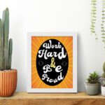 Work Hard Be Proud office wall art with white text on a black effect background gradient of orange and yellow, framed in white on the table. Perfect for home office decor.