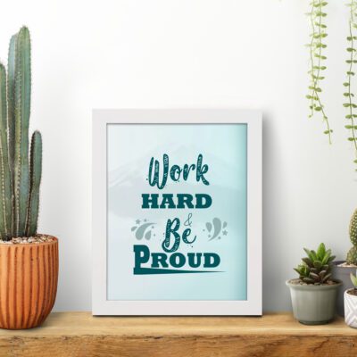 Work Hard Be Proud office wall art featuring green text with decorative elements on a white background, framed in white on the table. Perfect for home offices.