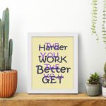 Infidu The Harder You Work The Better You Get wall art with a light sandal background dark blue and violet text and a black frame on the table. Inspiring decor for home offices and workspaces.