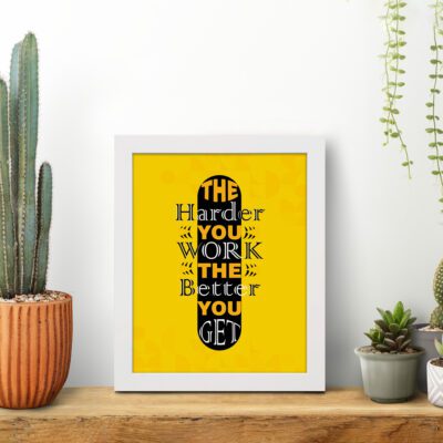 Infidu The Harder You Work The Better You Get wall art with a yellow background and dark yellow and white text with black shadow and white frame displayed on the table. Inspiring decor for home offices and workspaces.