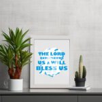 Wall art featuring the text The Lord Remembers Us And Will Bless Us in bold blue on a soft gray background. displayed on the table in a white frame