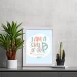 I Am a Child of God Christian Wall Art in bright orange and turquoise with a pastel blue background, perfect for home decor. displayed on the table in a white frame