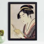 vintage art print features "Kobikicho Arayashiki Koiseya Ochie" by Utamaro Kitagawa, showing a traditional Japanese woman reading a book, mounted on the wall with black frame.