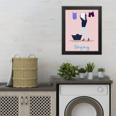 "Drying" Laundry Room Wall Art with a person hanging clothes on a line, light pink background, and blue laundry basket. hung on the wall the laundry wall art under the washing machine