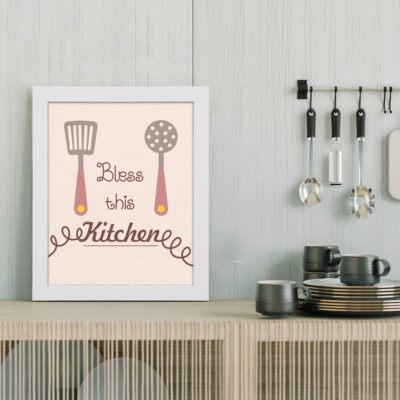 Bless This Kitchen wall art with spatula and ladle illustrations on a light pastel beige textured background. displayed on the table in a white frame