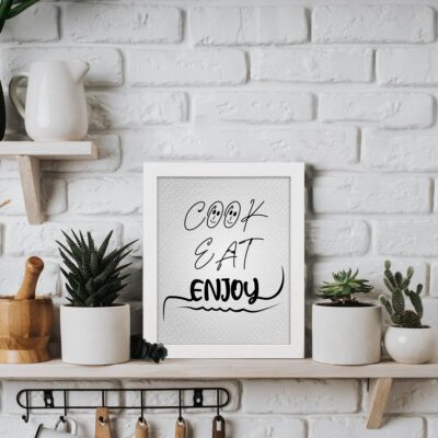 Cook Eat Enjoy kitchen wall art with black cursive text on a textured white background, featuring smiley faces above the word "Cook." displayed on the table in a white frame