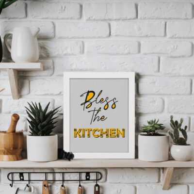 Infidu Bless This Kitchen Kitchen Wall Art with black and yellow fonts on a light gray background, modern kitchen decor piece. displayed on the table