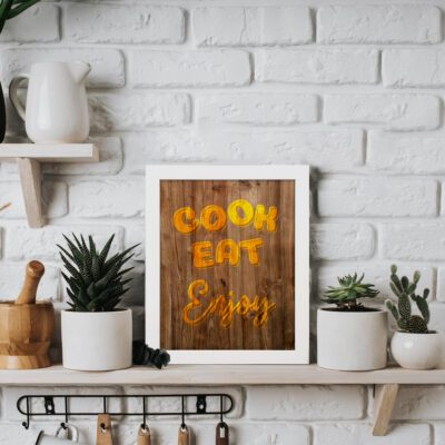 Cook Eat Enjoy kitchen wall art in glowing orange font on a rustic wooden background, adding warmth and charm to your kitchen decor. displayed on the table in a white frame