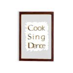 Cook Sing Dance kitchen wall art with black and orange text on a light gray background with jagged white accents. displayed on the wall in a brown frame in a white background