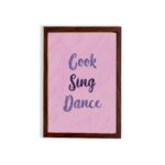 Infidu Cook Sing Dance Kitchen Wall Art with bold navy-blue text on soft pink background, framed in black, modern and lively kitchen design. displayed on the wall in a brown frame in a white background