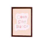 Cook Sing Dance Kitchen Wall Art with pastel pink, green, and blue text on a peach background, perfect for cheerful kitchen decor. displayed on the wall in a black frame in a brown background