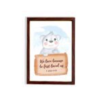 A cute white bear holding a wooden sign with the verse We Love Because He First Loved Us - 1 John 4:19 against a blue sky with clouds. hung on the wall in a brown frame