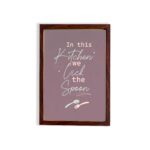 In This Kitchen We Lick The Spoon kitchen wall art with playful spoon illustration on a mauve background. displayed on the wall in a brown frame in a white background