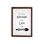 In This Kitchen We Lick the Spoon kitchen wall art featuring black text on a silver-gray background with a minimalist spoon illustration. displayed on the wall in a brown frame in a white background