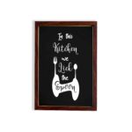 In This Kitchen We Lick The Spoon wall art with white cursive and bold fonts, black background, and stylish cutlery illustrations. displayed on the wall in a brown frame in a white background