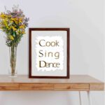 Cook Sing Dance kitchen wall art with black and orange text on a light gray background with jagged white accents.