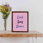 Infidu Cook Sing Dance Kitchen Wall Art with bold navy-blue text on soft pink background, framed in black, modern and lively kitchen design. displayed on the table