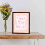 Cook Sing Dance Kitchen Wall Art with pastel pink, green, and blue text on a peach background, perfect for cheerful kitchen decor. displayed on the table