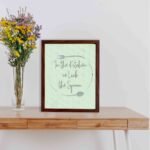 In This Kitchen We Lick The Spoon kitchen wall art with light green background and subtle kitchen utensil pattern in a casual handwritten font. displayed on the table