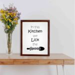 In This Kitchen We Lick the Spoon kitchen wall art featuring black text on a silver-gray background with a minimalist spoon illustration. displayed on the table