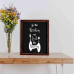 In This Kitchen We Lick The Spoon wall art with white cursive and bold fonts, black background, and stylish cutlery illustrations. displayed on the table