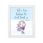 Wall art featuring a joyful bear in winter clothes holding a heart, with the verse We love because He first loved us from 1 John 4:19. hung on the wall in a white frame