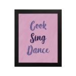 Infidu Cook Sing Dance Kitchen Wall Art with bold navy-blue text on soft pink background, framed in black, modern and lively kitchen design. displayed on the wall in a black frame in a white background