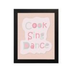 Cook Sing Dance Kitchen Wall Art with pastel pink, green, and blue text on a peach background, perfect for cheerful kitchen decor. displayed on the wall in a black frame in a black background