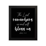 The Lord Remembers Us And Will Bless Us - Psalm 115:12 Bible Verse Wall Art on a black background with white text. hung on the wall in a black frame