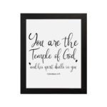 You Are The Temple Of God" Bible Wall Art with gold text on a black background from 1 Corinthians 3:16. hung on the wall in a black frame