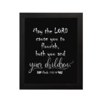 Wall art featuring the quote 'May the Lord cause you to flourish, both you and your children' from Psalm 115:14 on a black background. hung on the wall in a black frame