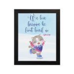 Wall art featuring a joyful bear in winter clothes holding a heart, with the verse We love because He first loved us from 1 John 4:19. hung on the wall in a black frame