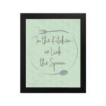 In This Kitchen We Lick The Spoon kitchen wall art with light green background and subtle kitchen utensil pattern in a casual handwritten font. displayed on the wall in a black frame in a white background