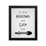 In This Kitchen We Lick the Spoon kitchen wall art featuring black text on a silver-gray background with a minimalist spoon illustration. displayed on the wall in a black frame in a white background