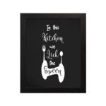 In This Kitchen We Lick The Spoon wall art with white cursive and bold fonts, black background, and stylish cutlery illustrations. displayed on the wall in a black frame in a white background