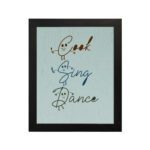 Cook Sing Dance kitchen wall art with hand-drawn fonts, colorful doodles, and a textured light blue background. displayed on the wall in a black frame in a white background