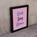 Infidu Cook Sing Dance Kitchen Wall Art with bold navy-blue text on soft pink background, framed in black, modern and lively kitchen design. displayed on the wall in a black frame